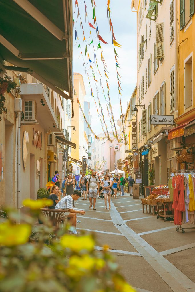 Bustling street in Ajaccio, France with shops, cafes, and pedestrians under vibrant flags. Rue animée d'Ajaccio, France avec des boutiques, des cafés et des piétons sous des drapeaux vibrants.