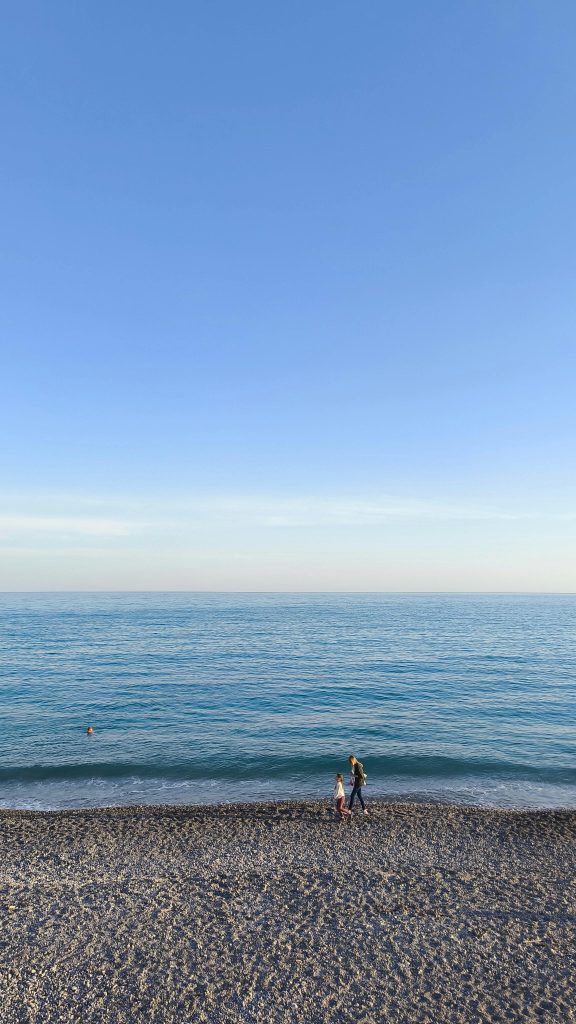 A serene scene of a mother and daughter strolling on the beach in Nice, France, under a clear blue sky. Une scène sereine d'une mère et sa fille se promenant sur la plage de Nice, en France, sous un ciel bleu clair.
