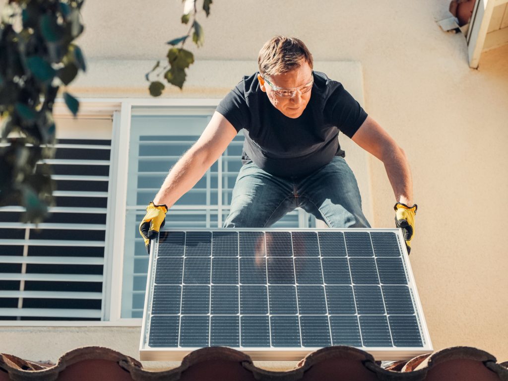 A man in safety glasses installs a solar panel on a house roof, promoting renewable energy. Un homme portant des lunettes de sécurité installe un panneau solaire sur le toit d'une maison, promouvant l'énergie renouvelable.