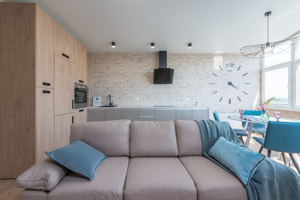 Minimalistic interior design of modern light studio consisting of big sofa and kitchen with dining table and chairs against brick wall in daylight Design d'intérieur minimaliste d'un studio de lumière moderne composé d'un grand canapé et d'une cuisine avec table à manger et chaises contre un mur de briques à la lumière du jour